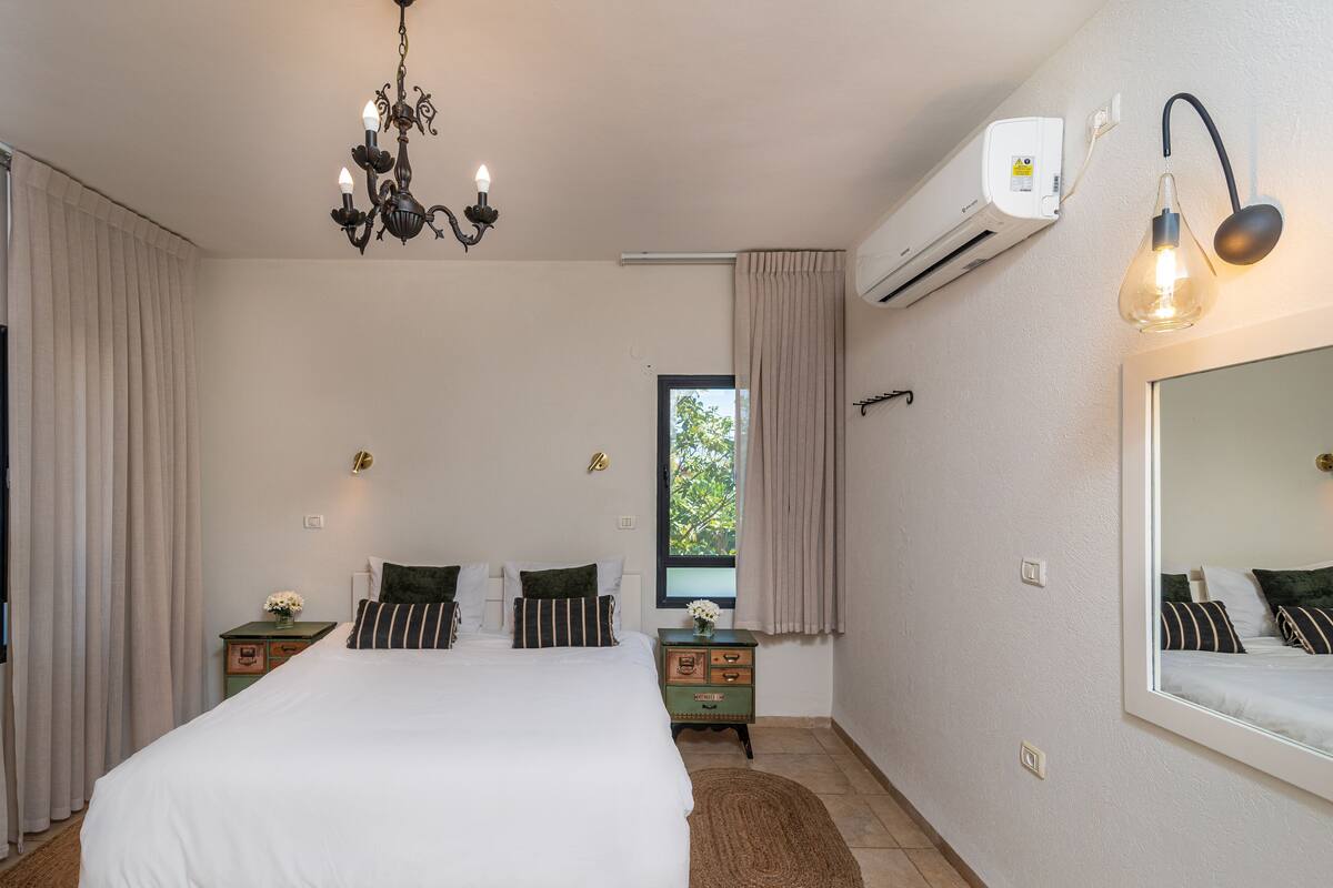A bedroom with a bed, a chandelier, an air conditioning unit, a mirror, and a window.