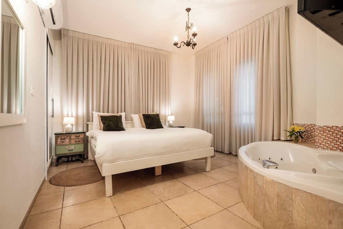 A bedroom with a large bed, a bedside table with a lamp, a bathroom with a bathtub, and a chandelier.