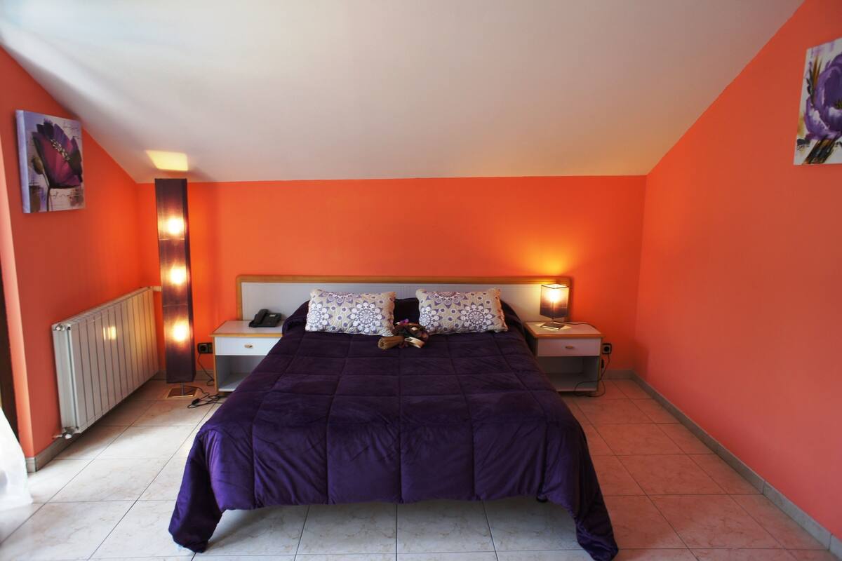 A bedroom with a purple bedspread, two bedside tables with lamps, a radiator, and artwork on the walls.
