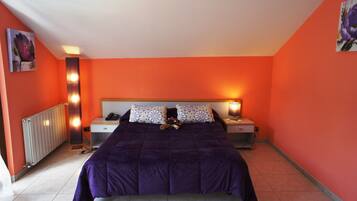 A bedroom with a purple bedspread, two bedside tables with lamps, a radiator, and artwork on the walls.