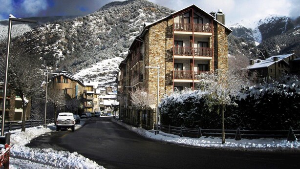 A snow-covered mountain town with a stone building featuring multiple balconies and a ski slope in the background. - Hotel la Planada (Ordino)