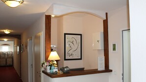 A reception area with a wooden reception desk, a framed quote on the wall, a lamp, and a small vase with flowers. - Hotel Gardenia (Reiskirchen)