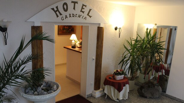 A hotel entrance with a sign reading "HOTEL GARDENIA," a reception desk with a lamp, and potted plants. - Hotel Gardenia (Reiskirchen)