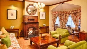 Un salón tradicional con chimenea, sillones verdes y cortinas con motivos florales.