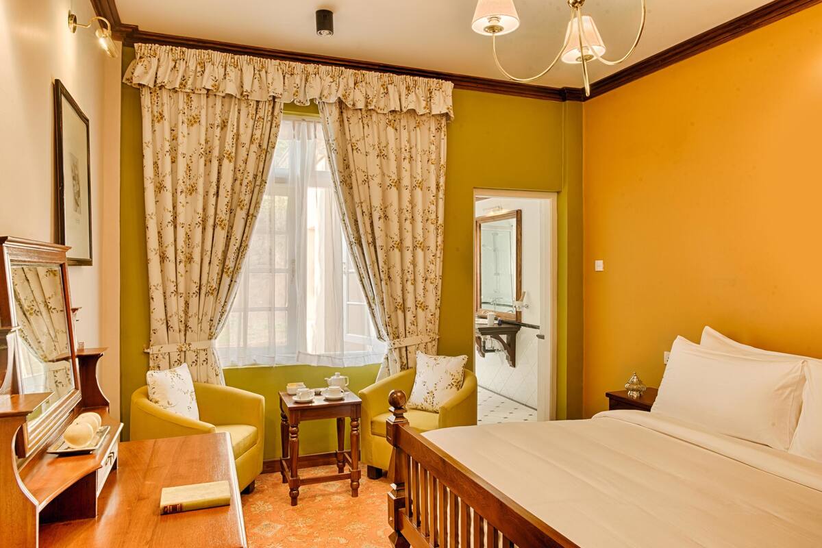 Habitación de hotel con cama, escritorio, silla, una mesita con lámpara y ventana con cortinas.