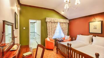 A bedroom with a large bed, a wooden desk, a chair, and a small table. There is a window with curtains and a door leading to another room.
