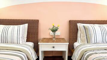 A hotel room with two beds, a nightstand with a vase of flowers, and a plain wall.