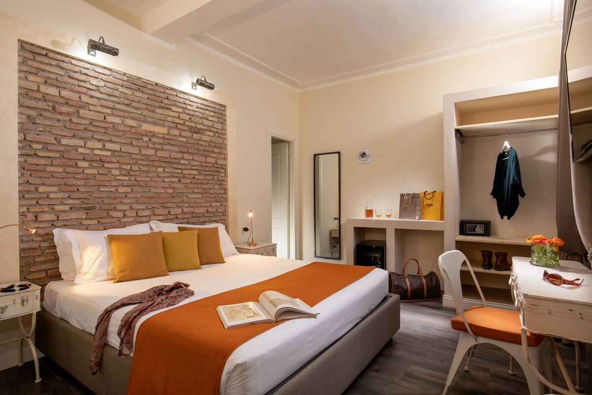 A hotel room with a brick accent wall, a large bed with an orange throw, a desk with a chair, and a mirror.