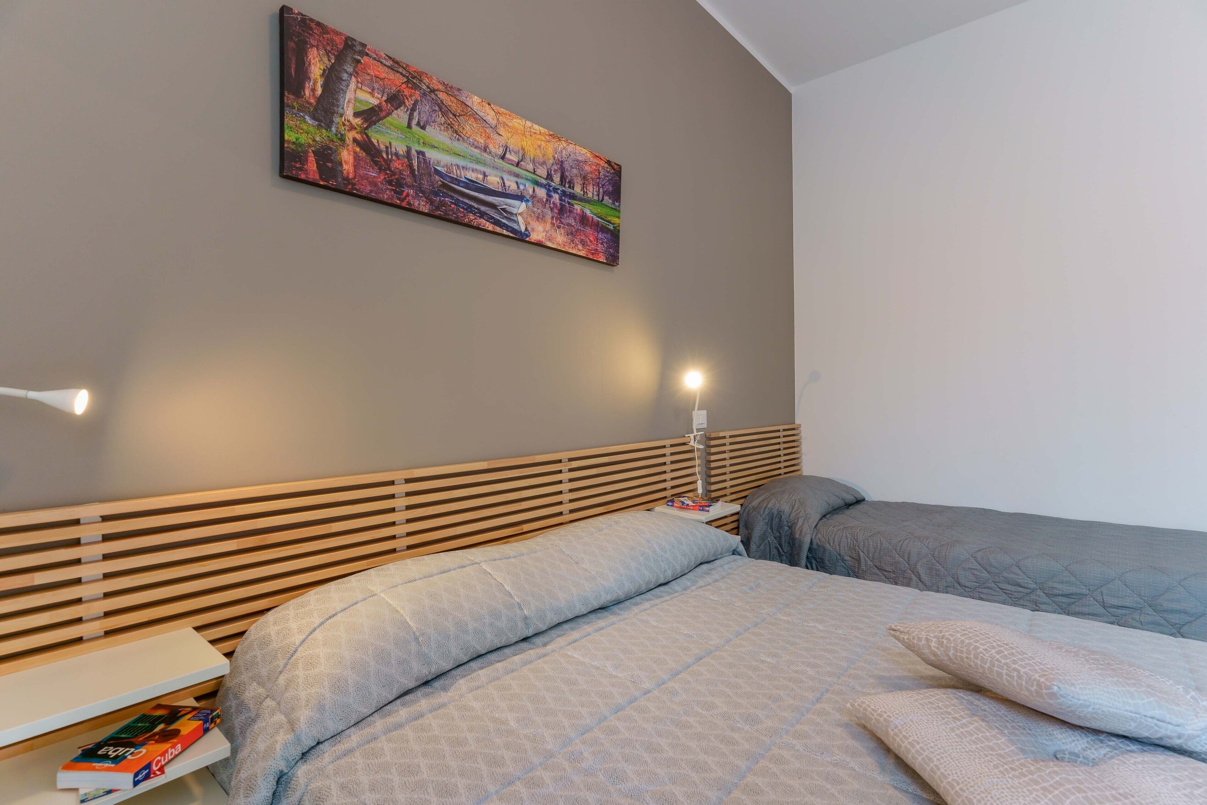 A bedroom with a wooden slat headboard, a bed with a quilted cover, a bedside table with books, and a painting on the wall.