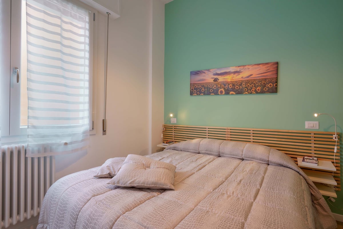 Una camera da letto con un letto, un termosifone, una finestra con persiane, un quadro appeso al muro e un comodino con una lampada.