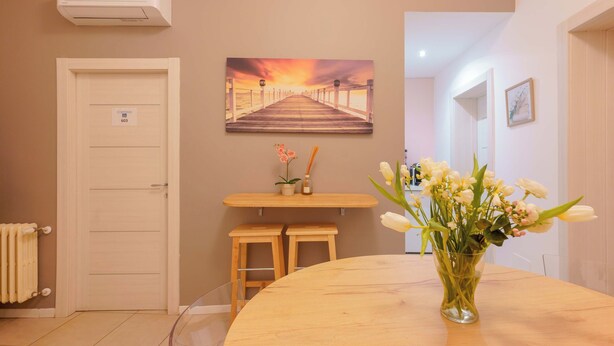 A hotel room with a wooden table, a vase of flowers, a painting of a pier, and a door with a sign. - B&B Bentivogli (Bologna)