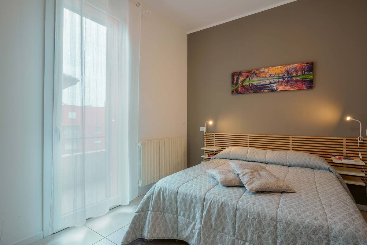 Una camera da letto con un letto, una finestra con tende leggere, un termosifone, una testiera e un quadro appeso al muro.