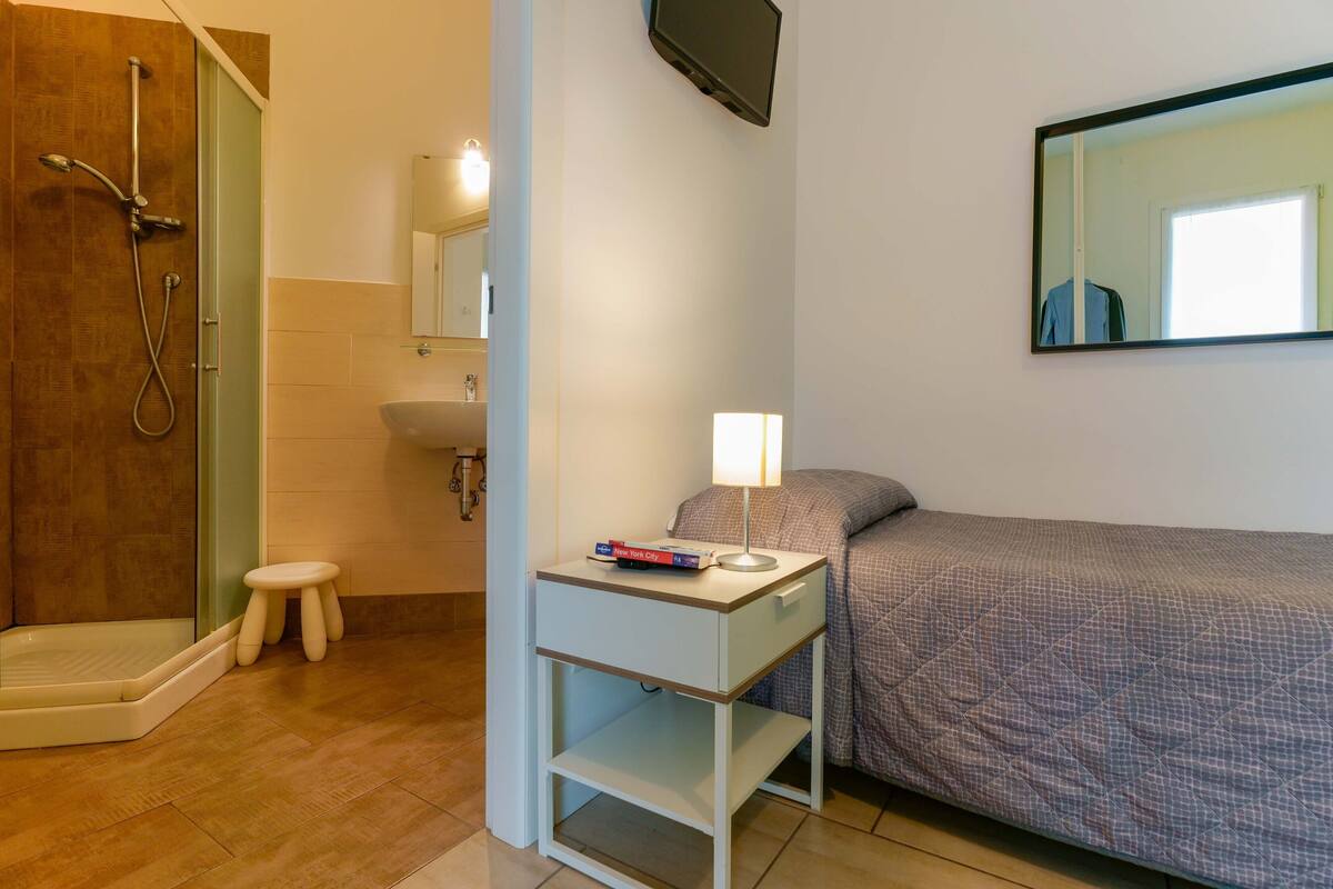 Un bagno con doccia, toilette e lavandino. È presente un comodino con lampada e libri.