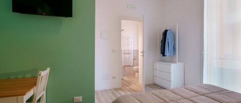 A bedroom with a bed, a TV mounted on the wall, a wooden table with a chair, and a bathroom visible through an open door.