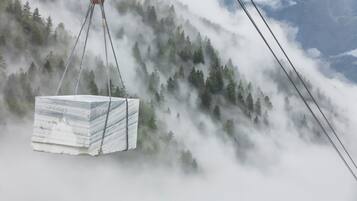 Un bloque grande de mármol blanco siendo levantado por una grúa frente a un paisaje montañoso cubierto de bosque.