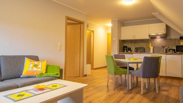 A compact living space with a sofa, dining table, and chairs, a kitchenette, and a wooden door. - Pension & FeWo's Talblick (Eibenstock)