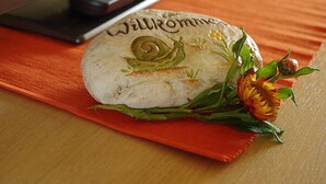 A welcome sign with a snail and flowers on a table with a remote control in the background. - Pension & FeWo's Talblick (Eibenstock)