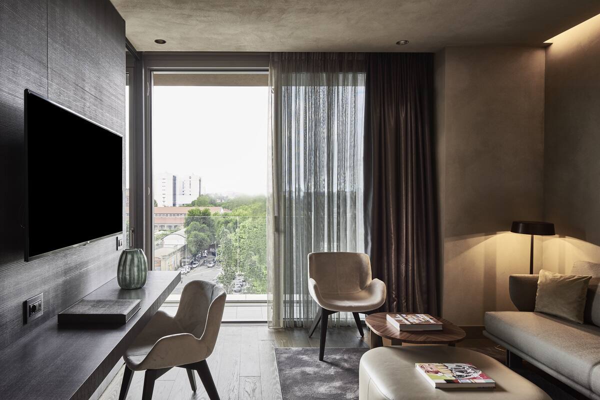 Una camera d'albergo moderna con una grande televisione a schermo piatto, un divano, una poltrona, un tavolino da caffè e vista sulla città attraverso ampie finestre.