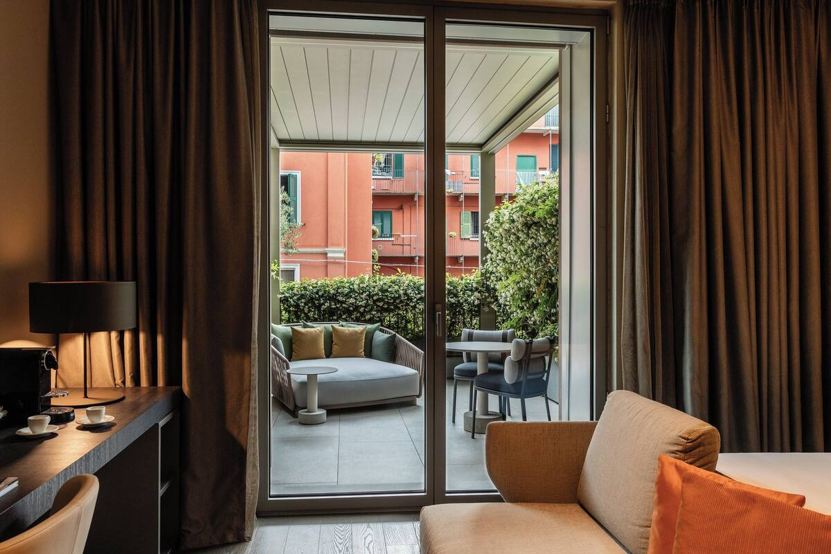 Una camera d'albergo con divano, un tavolino, una lampada e vista su un balcone esterno con tavolo e sedie.