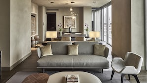 Una sala de estar moderna con un sofá gris, un sillón blanco y una mesa de centro. Hay un comedor con una mesa y sillas, y un ventanal que ofrece vistas de la ciudad.