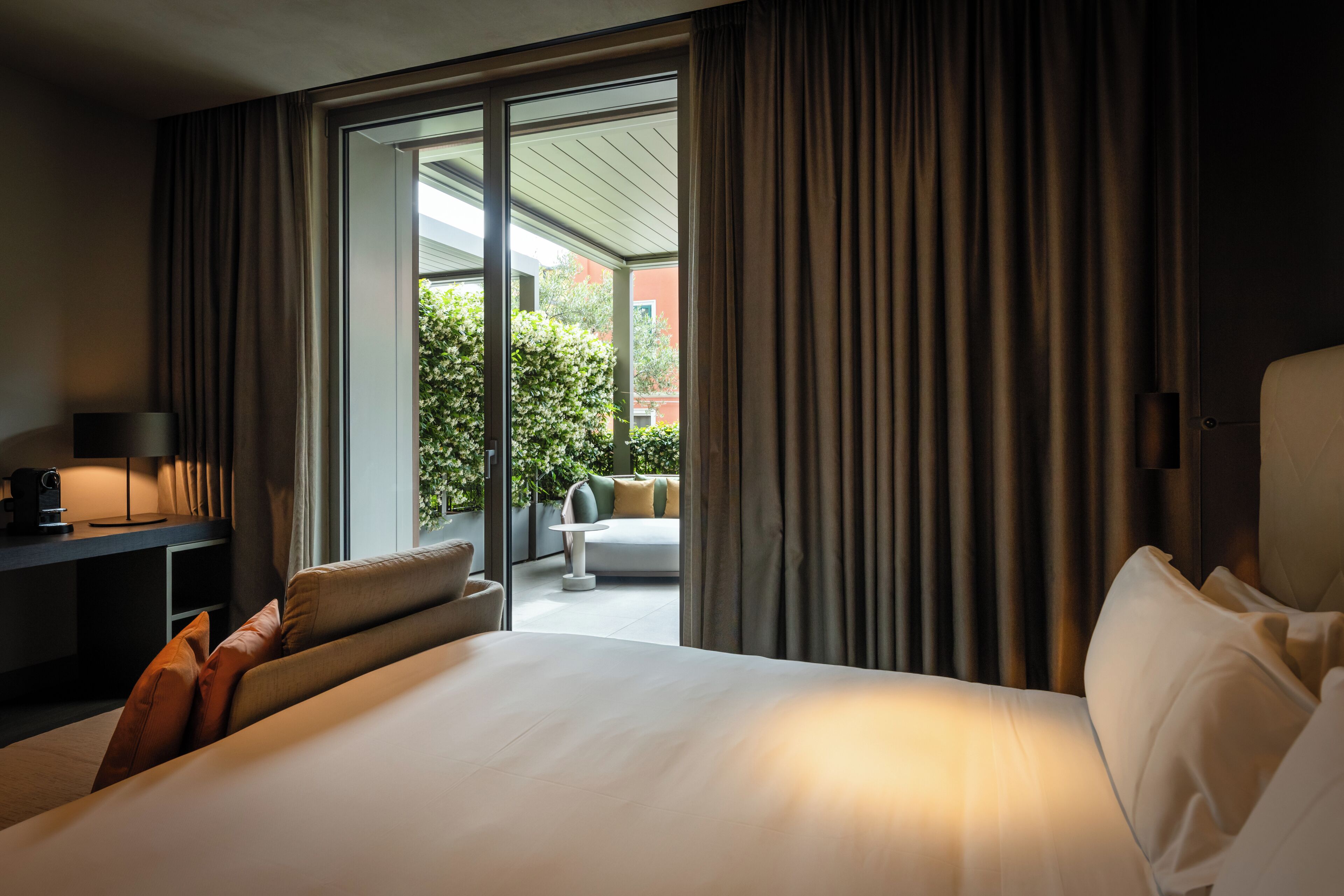 Camera d'albergo con un letto grande, una scrivania con una lampada e vista su un balcone con divano e piante.