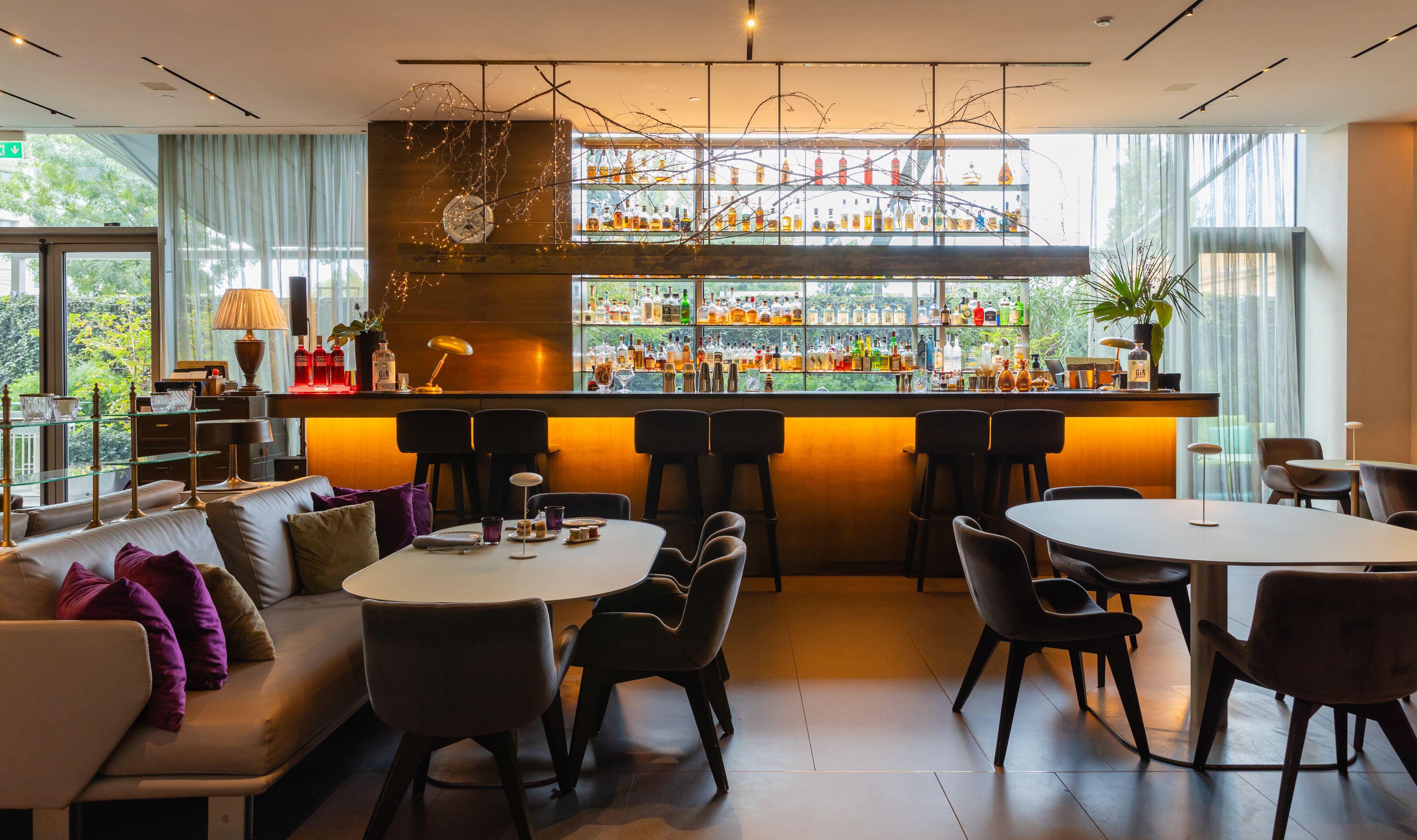 Un espace bar moderne avec un assortiment de bouteilles d’alcool, un coin salon doté de canapés et de fauteuils, et de grandes fenêtres offrant une vue sur la verdure environnante.