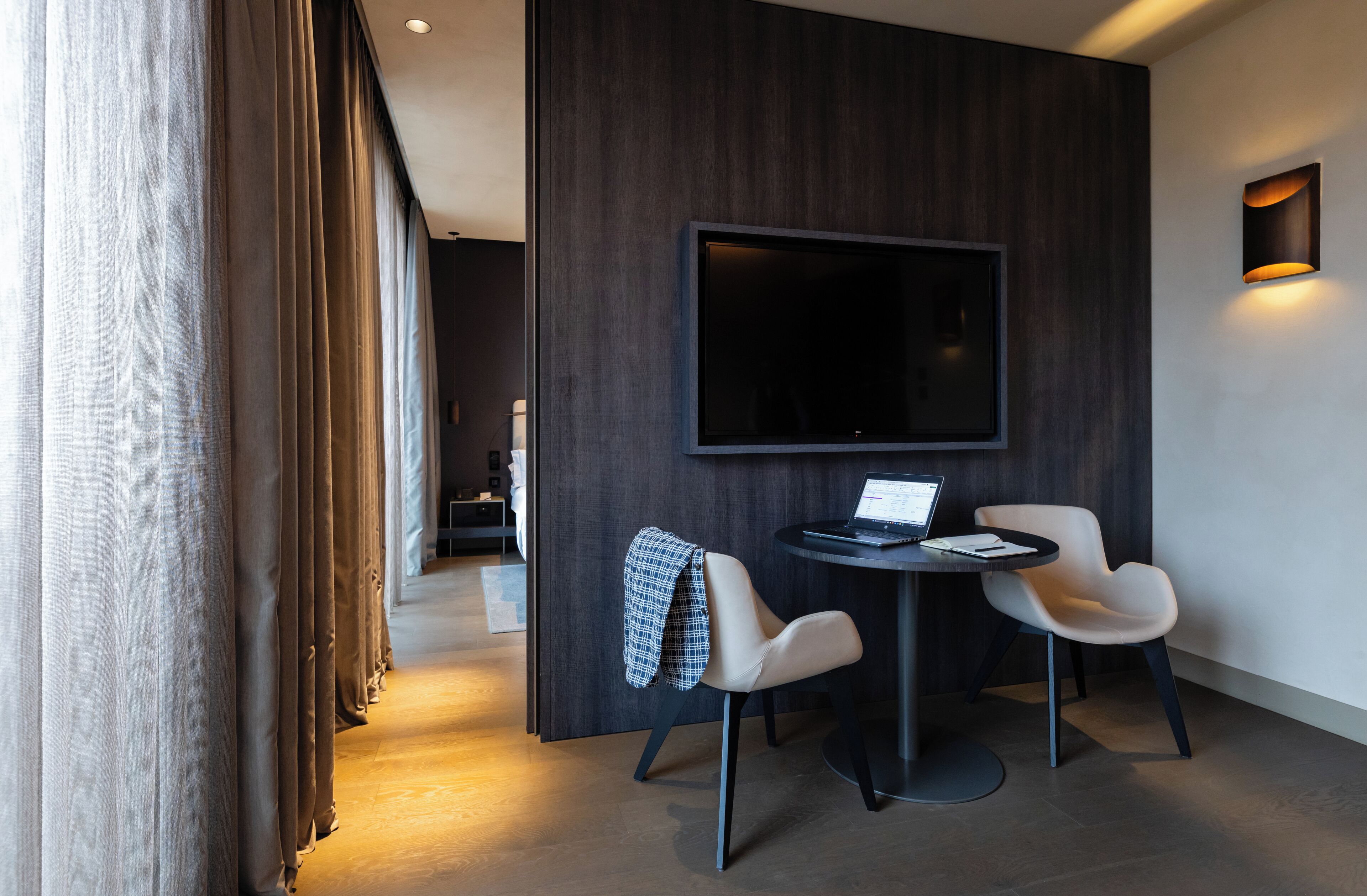 Une chambre moderne dotée d’un grand téléviseur à écran plat fixé au mur, de deux fauteuils disposés autour d’une petite table et d’un ordinateur portable posé sur la table.