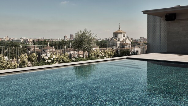 Una piscina en la azotea con agua cristalina, rodeada por una cerca metálica, con vistas a un paisaje urbano con edificios y una iglesia.