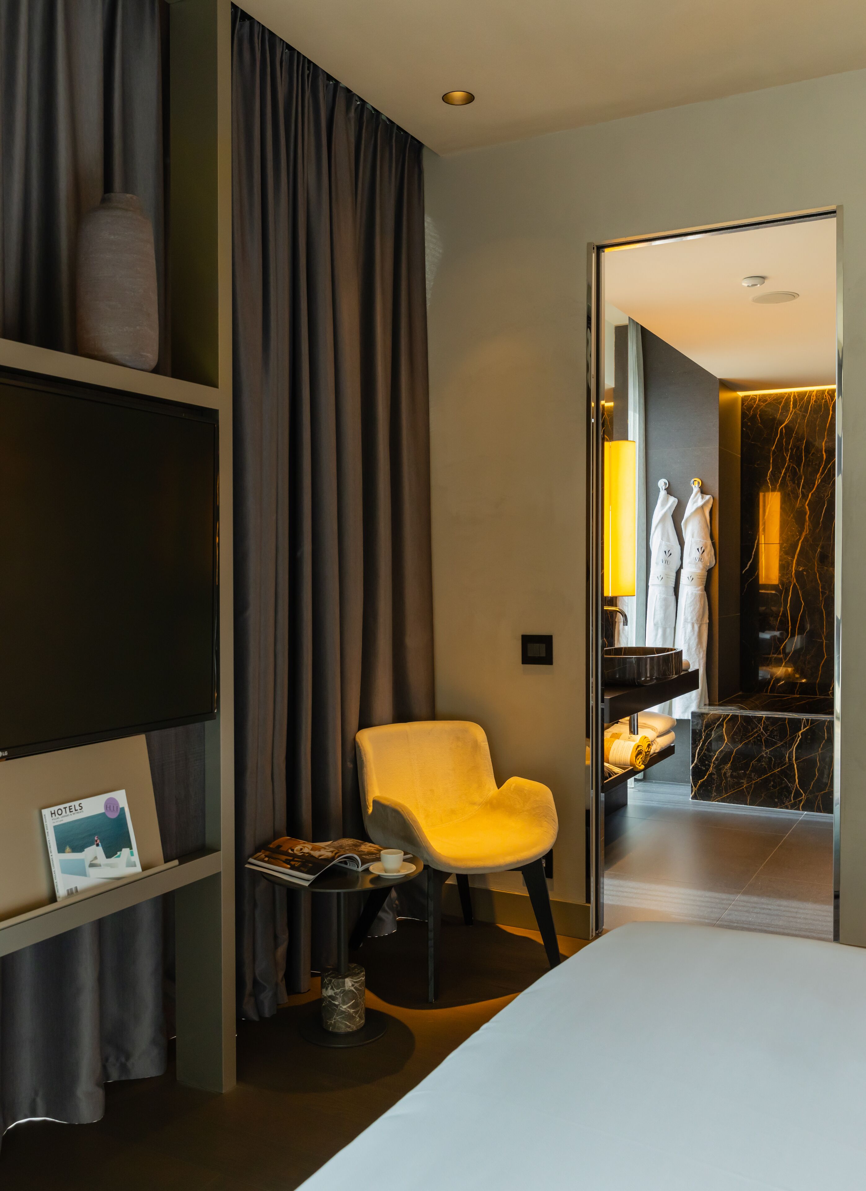 Habitación de hotel con una cama, un sillón amarillo, una mesita con una revista, una estantería con un florero, y un baño visible a través de una puerta abierta.