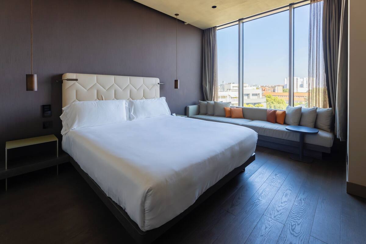 Camera d'albergo moderna con un letto grande, una panca con cuscini, un tavolino e un'ampia finestra che si affaccia sulla città.