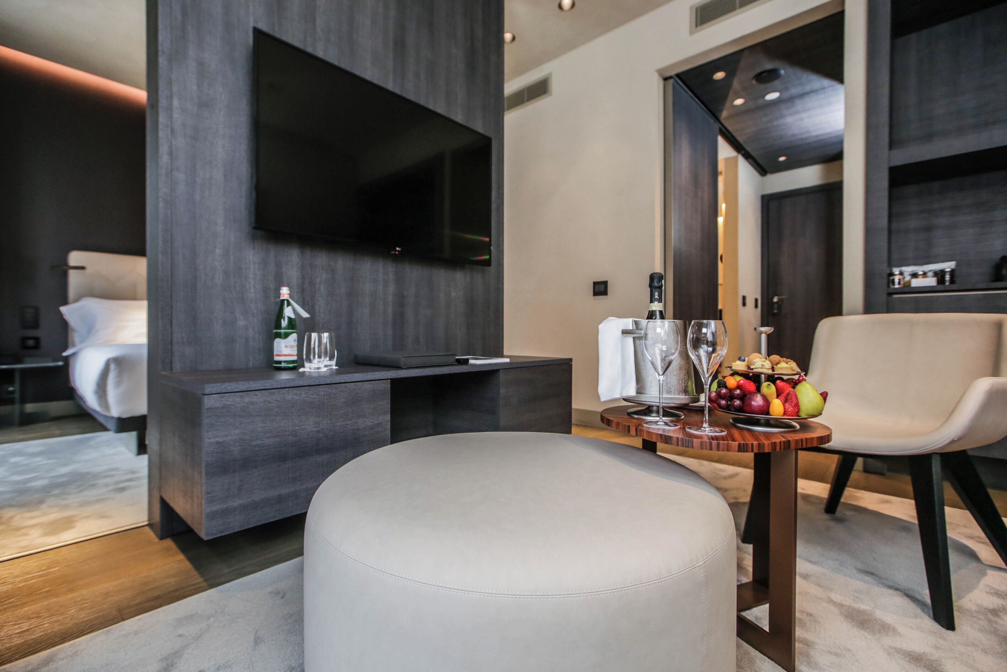 Una habitación de hotel moderna con una televisión de pantalla plana montada en la pared, un minibar, una zona de estar con un taburete redondo y una mesa de comedor con una bandeja de frutas.