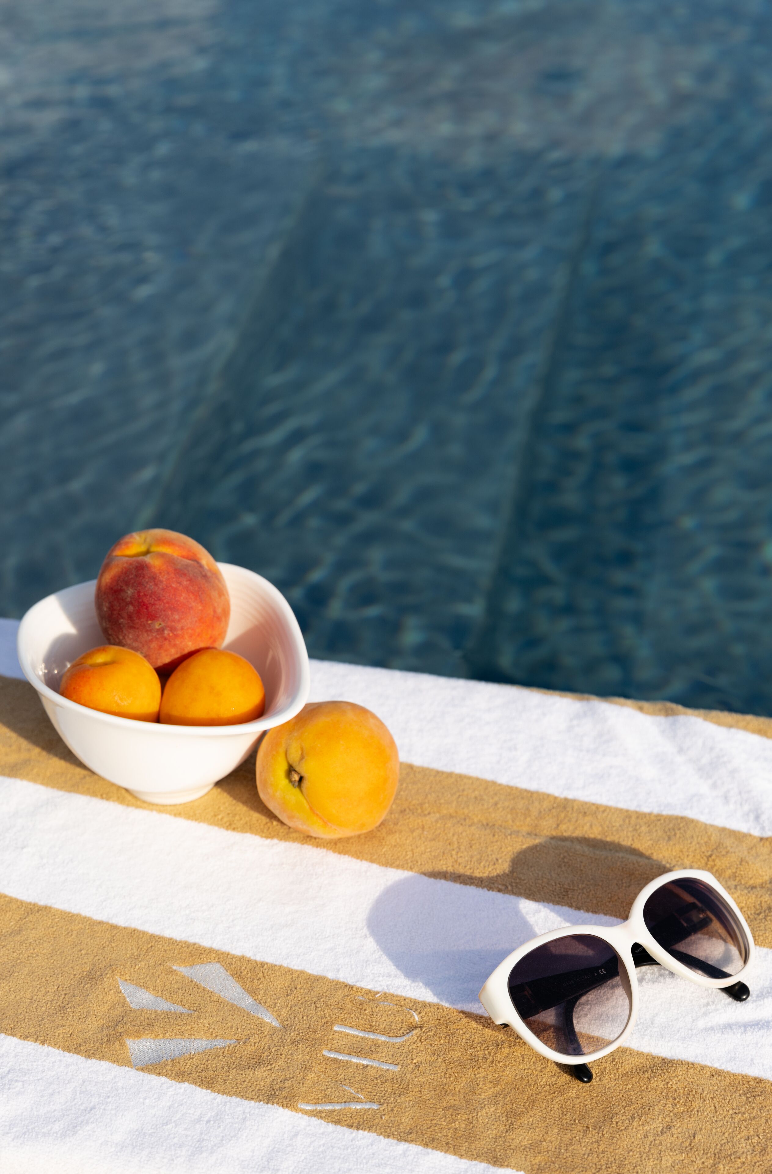 Una ciotola di frutta e occhiali da sole su un asciugamano a righe vicino a una piscina.