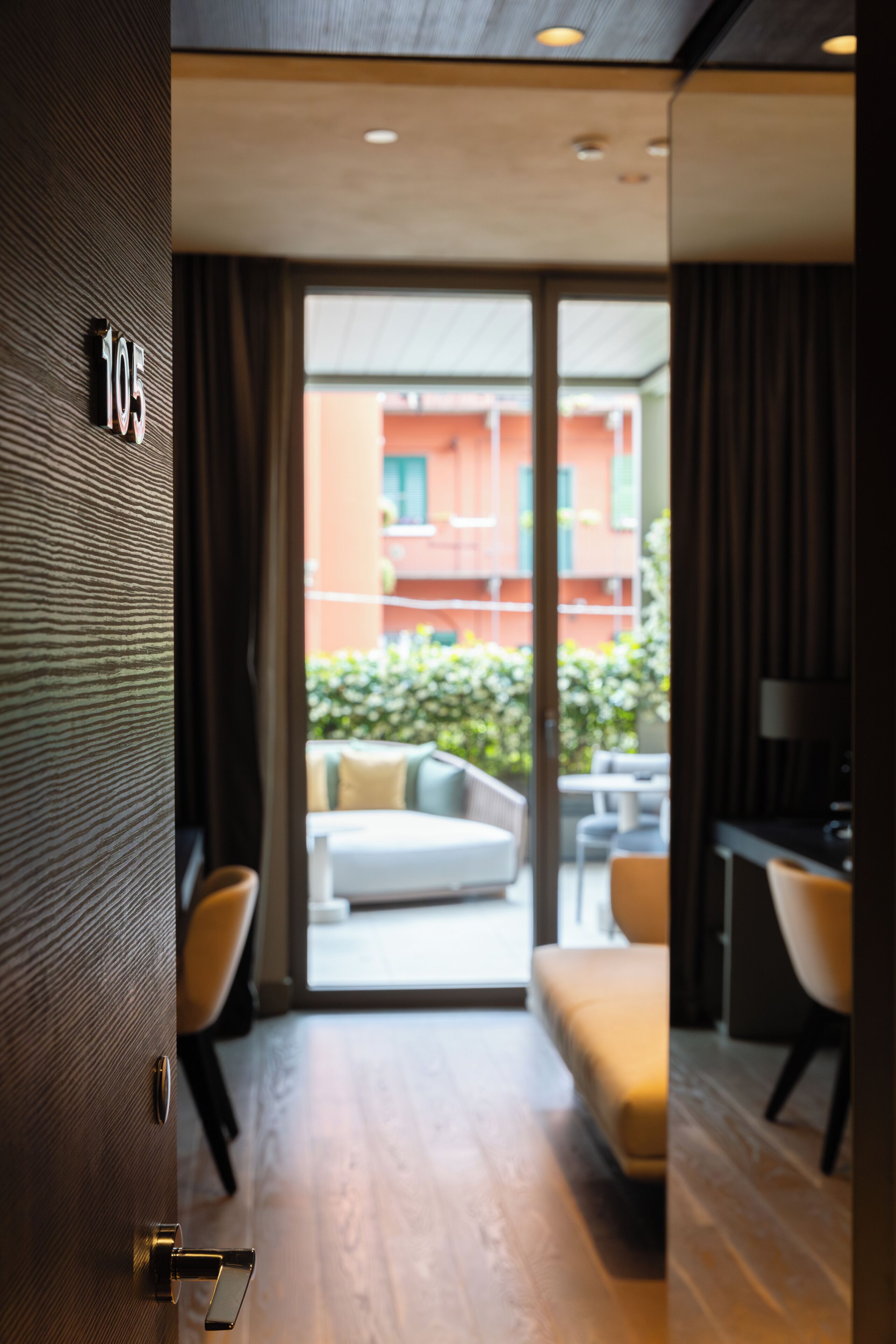 A modern hotel room with a wooden floor, a sofa, a desk, and a view of the outside balcony.