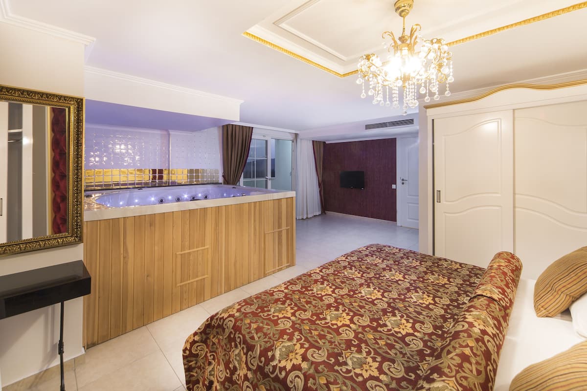 A bedroom with a large bed, a vanity with a mirror, a built-in bathtub, and a chandelier.