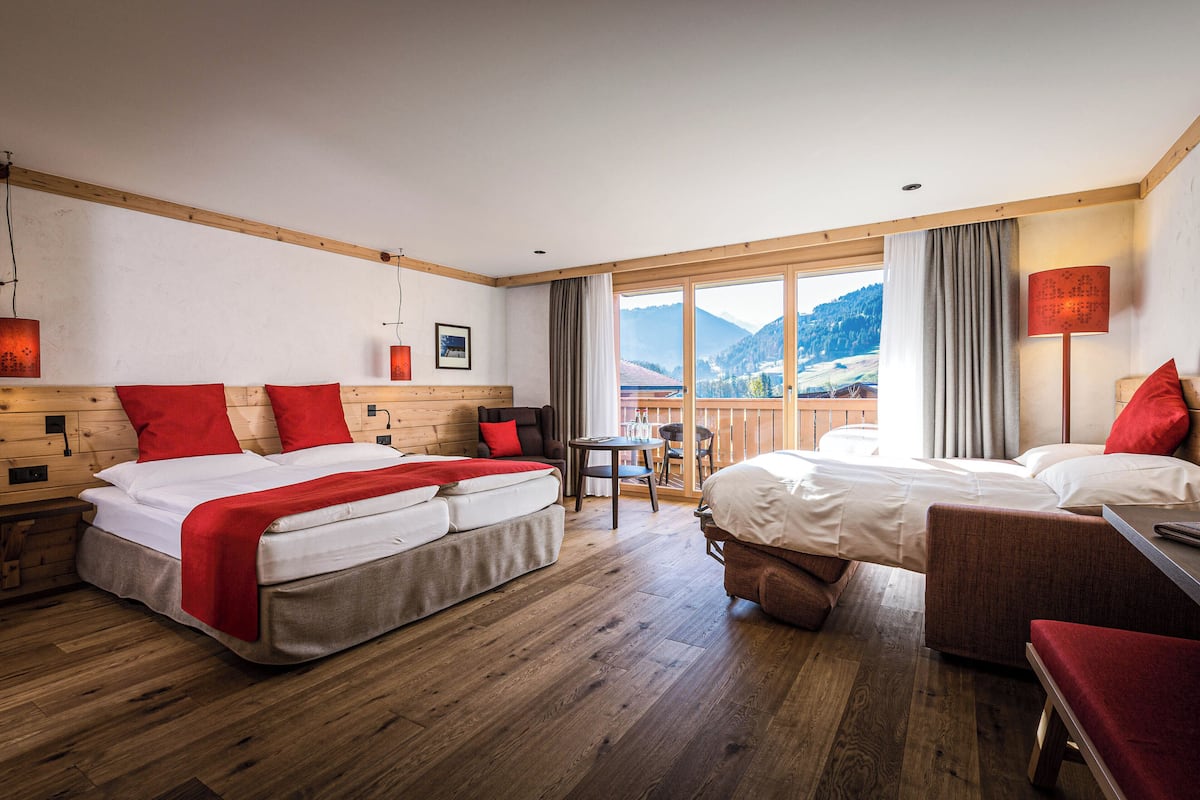 A hotel room with a large bed, a smaller bed, a sofa, and a table. There is a view of mountains through the window.