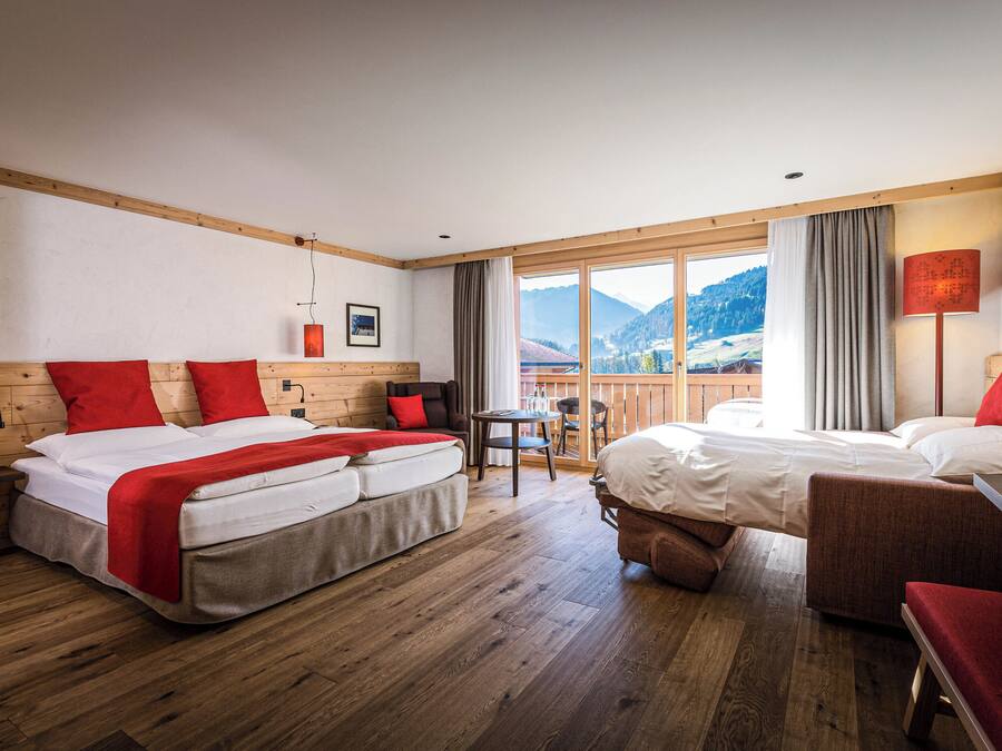 Une chambre d’hôtel avec un grand lit, un plus petit lit, un canapé et une table. La fenêtre offre une vue sur les montagnes.