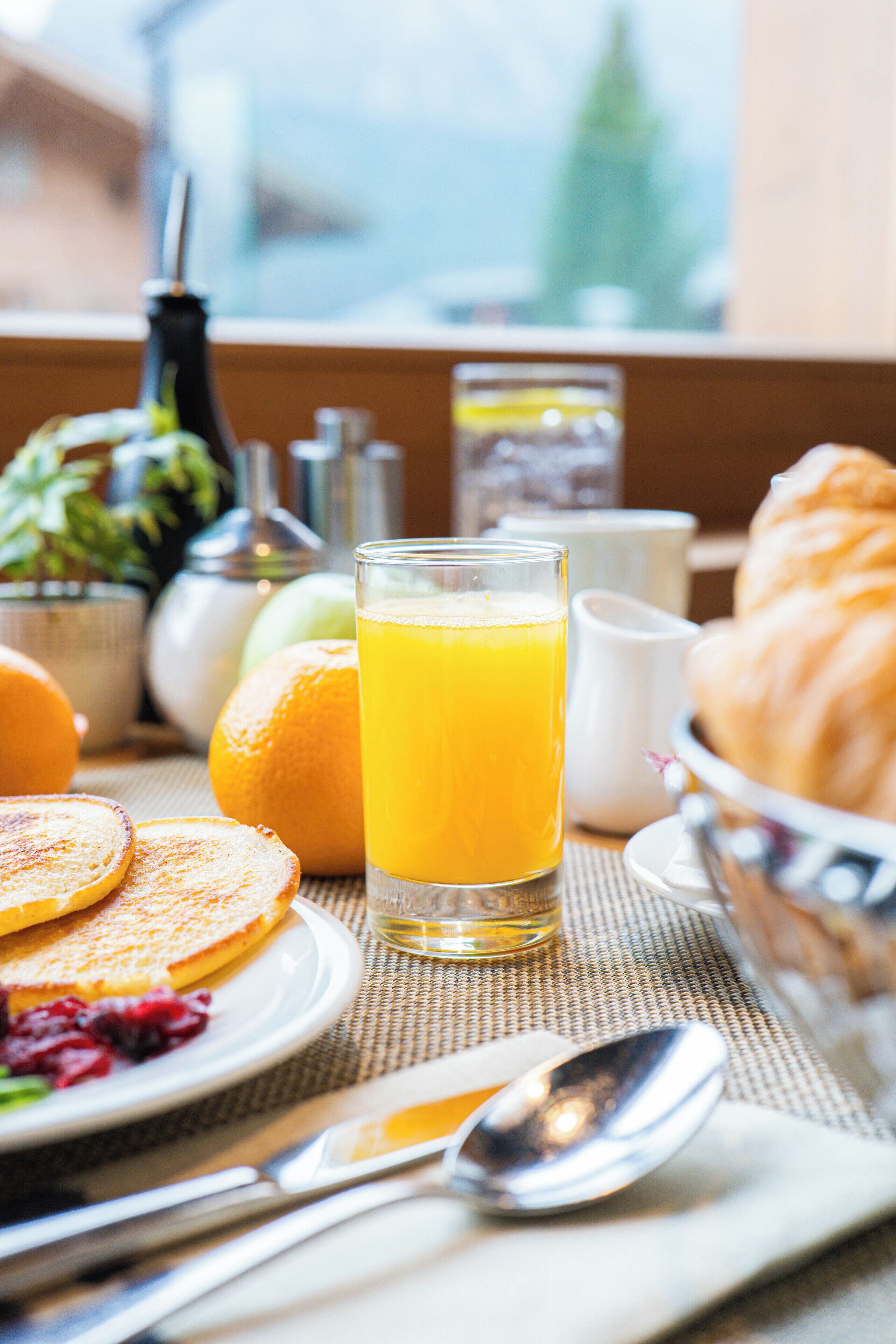 Desayuno con tortitas, zumo de naranja, cruasanes y un bol de fruta.