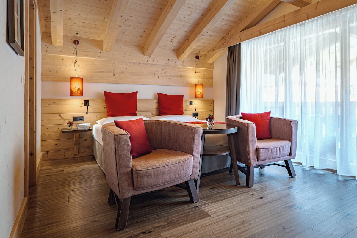 A wooden-paneled room with two sofas and a table, large windows with sheer curtains, and wall-mounted lamps.