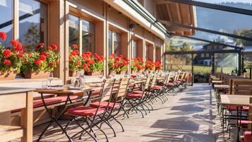 An outdoor café with rows of tables and chairs, potted red flowers, and a view of the surrounding landscape.