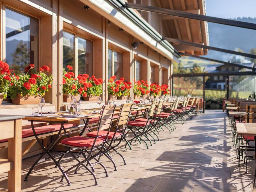 An outdoor café with rows of tables and chairs, potted red flowers, and a view of the surrounding landscape.