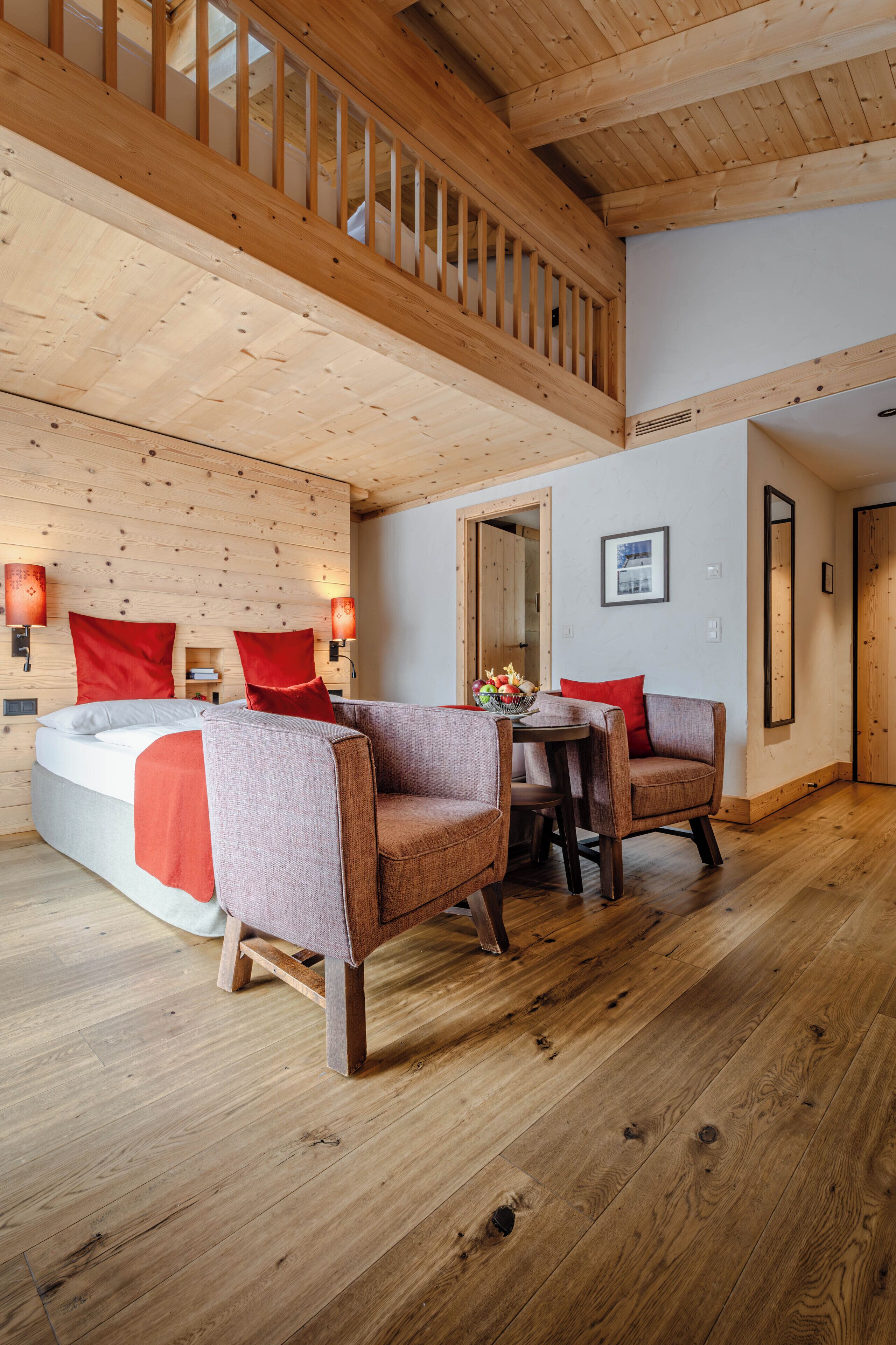 Una habitación con suelo de madera, una cama, dos sillones, una mesa y una escalera.