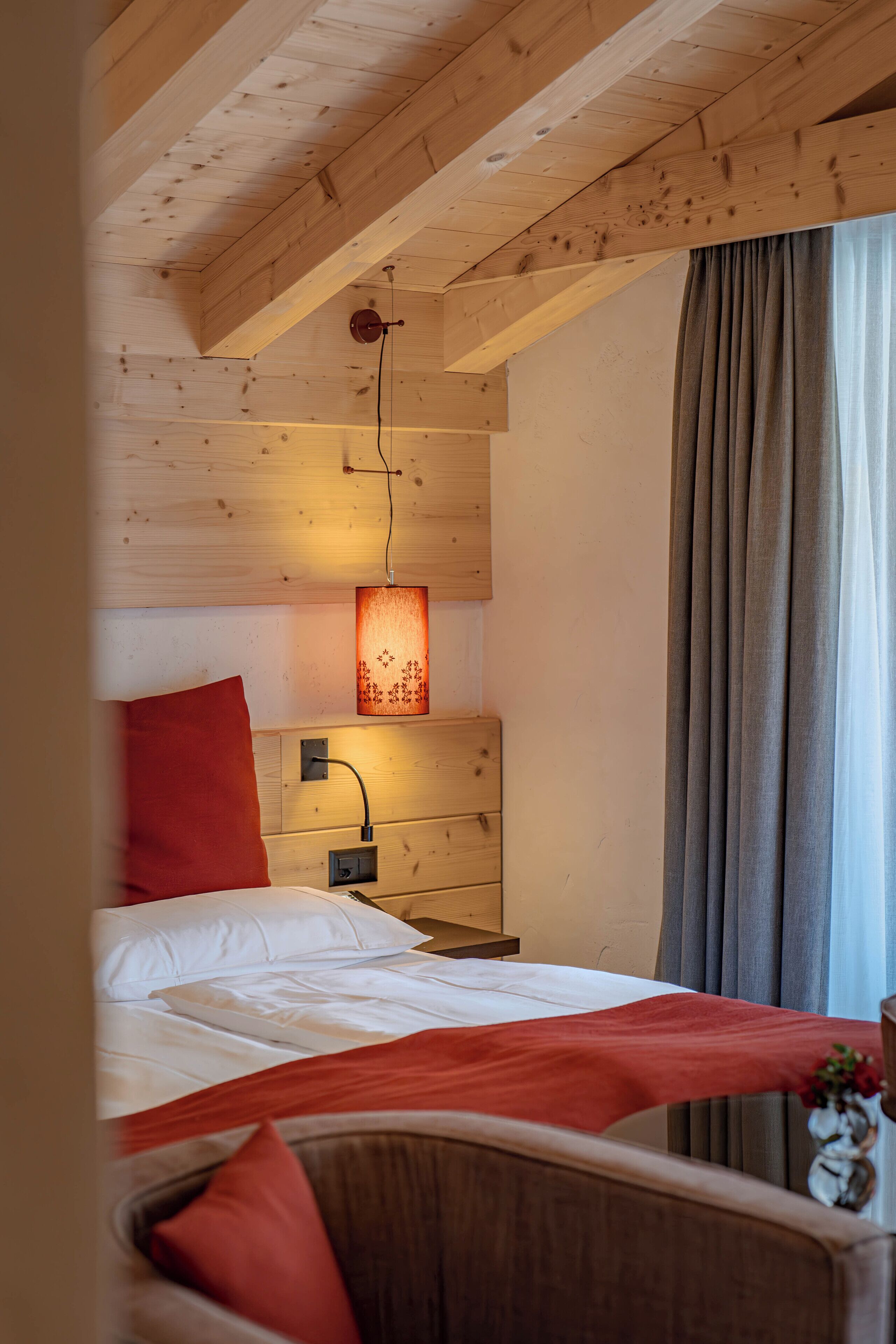 A wooden ceiling with exposed beams, a bed with white linens and a red accent pillow, a bedside table with a lamp, and a window with curtains.