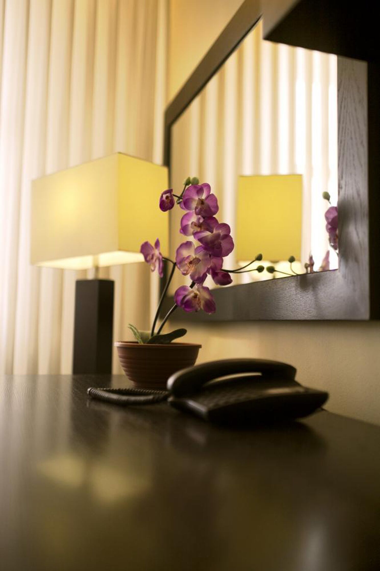 A hotel room with a bedside table, a lamp, and a mirror reflecting the room's interior.