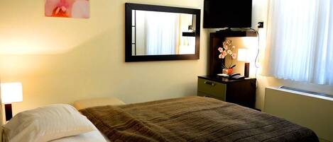 Quarto de hotel com cama, mesa de cabeceira, escrivaninha com luminária, televisão e janela com cortinas.