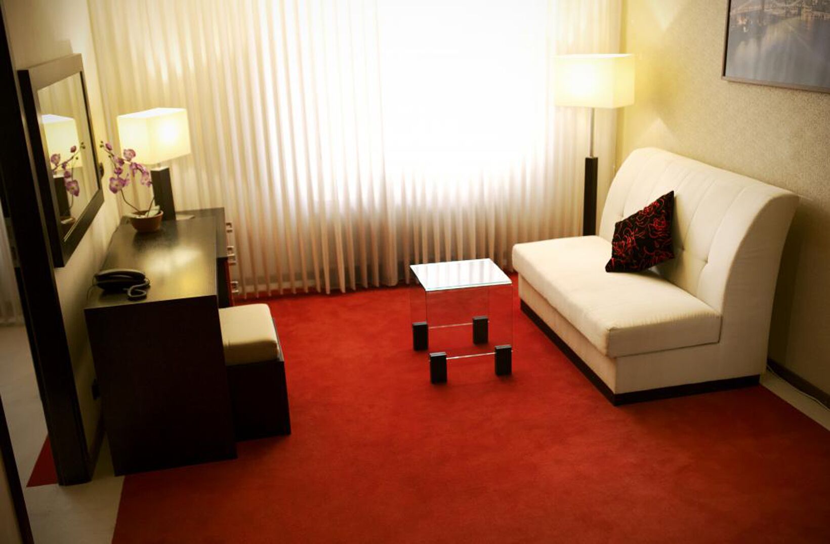 A hotel room with a white sofa, a small coffee table, a desk with a lamp, and a red carpet.