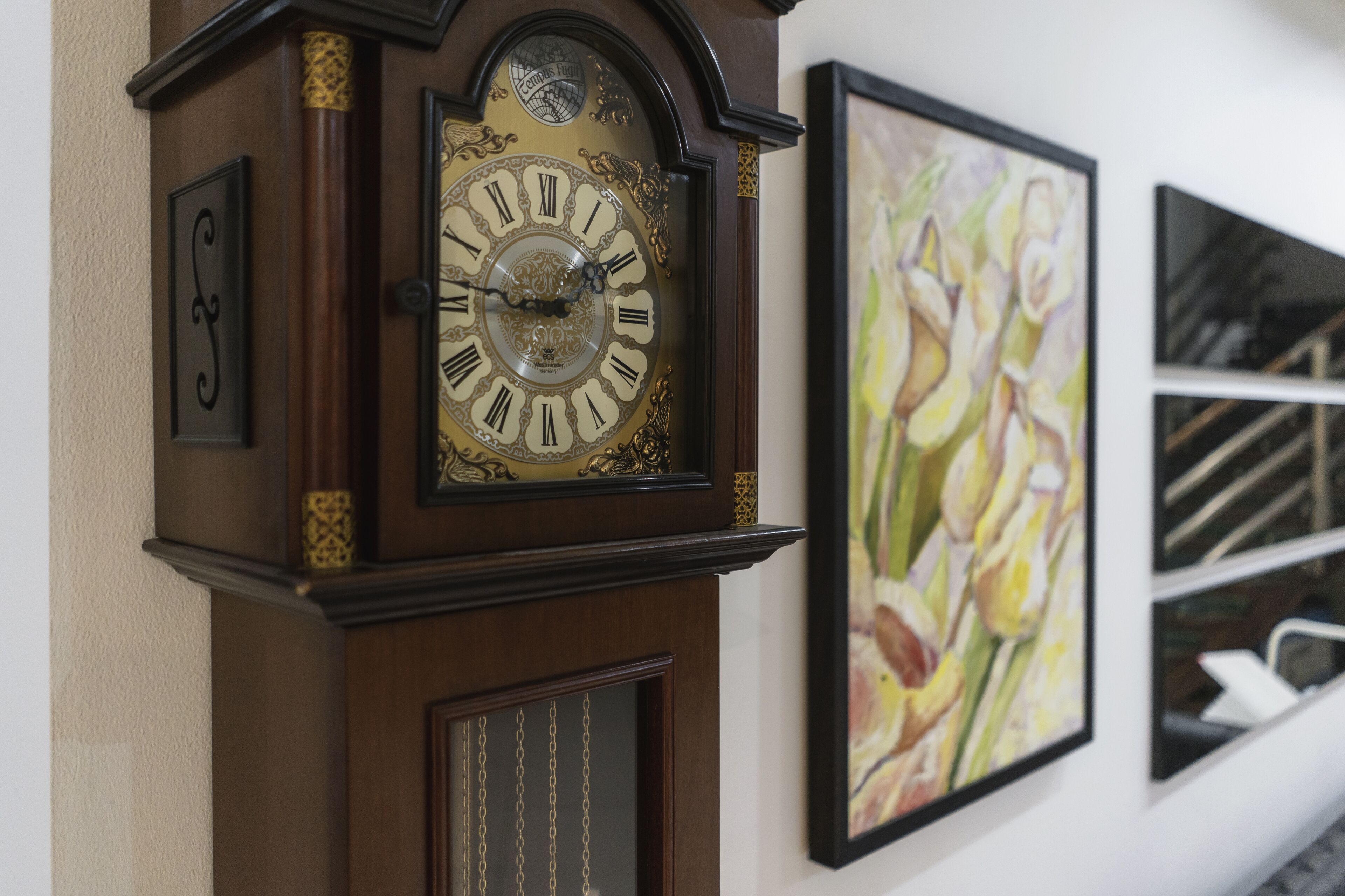 Un reloj de pared con números romanos y un péndulo, junto a un cuadro enmarco con motivos florales.
