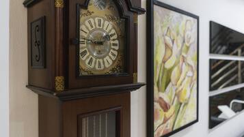 Un reloj de pared con números romanos y un péndulo, junto a un cuadro enmarco con motivos florales.