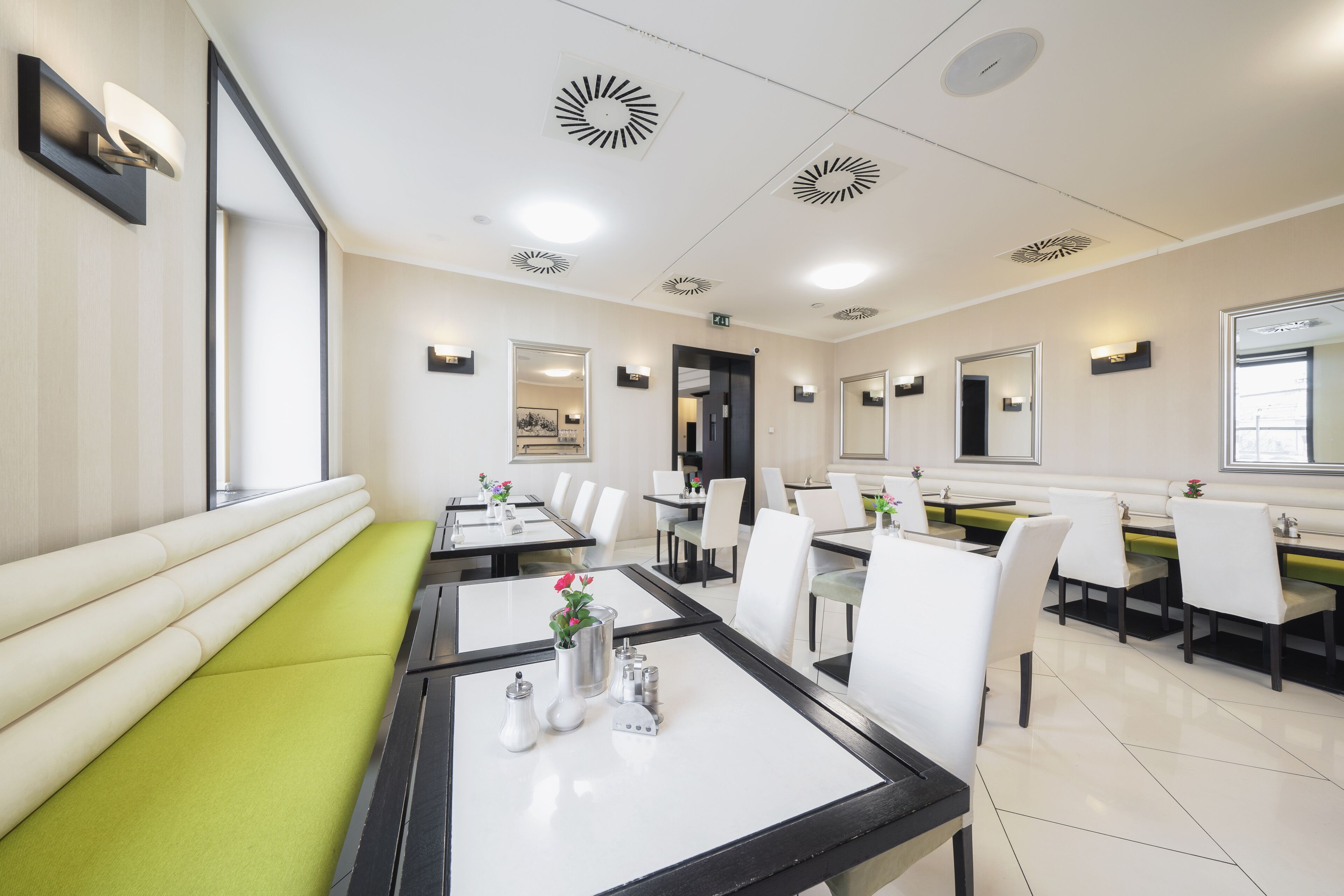 A modern dining area with multiple tables and chairs, a green cushioned bench, and ceiling vents.