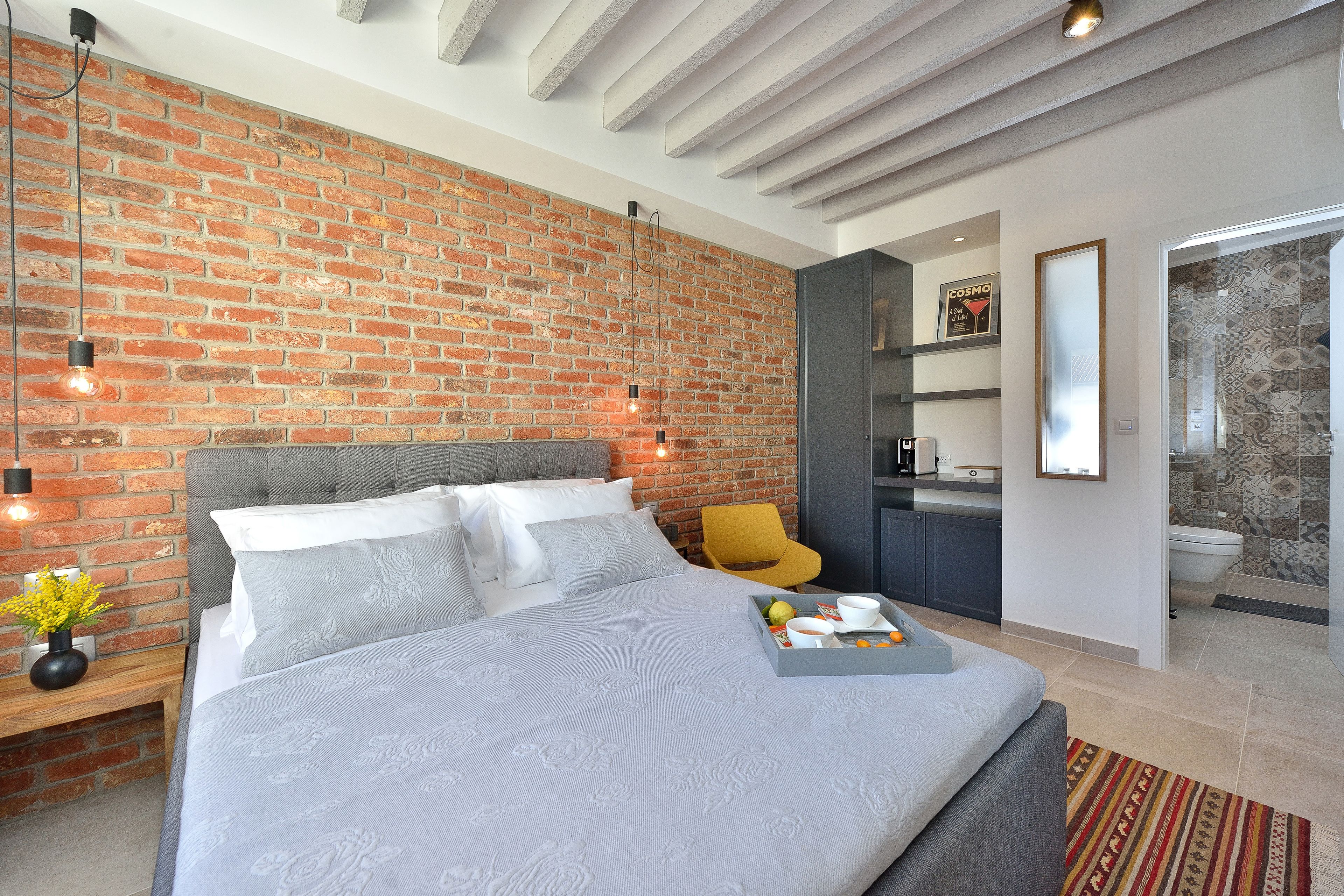 Un dormitorio moderno con pared de ladrillo, un cabecero tapizado en gris y una mesita de noche con una lámpara y flores.