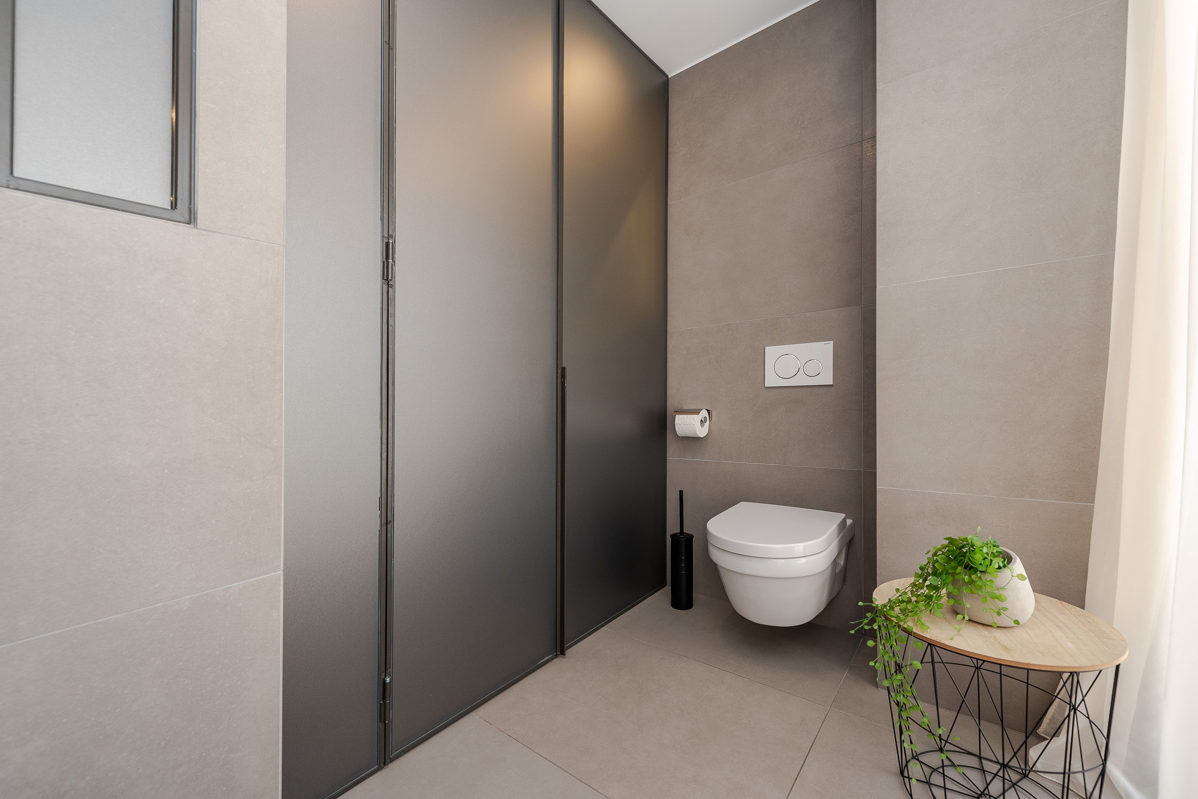 Une salle de bain moderne équipée de toilettes, d’un bidet, d’un porte-rouleau de papier toilette et d’une petite table ronde agrémentée d’une plante.